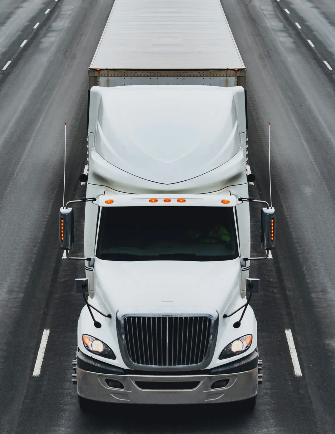 A white semi-truck with its headlights on, driving down a multi-lane highway