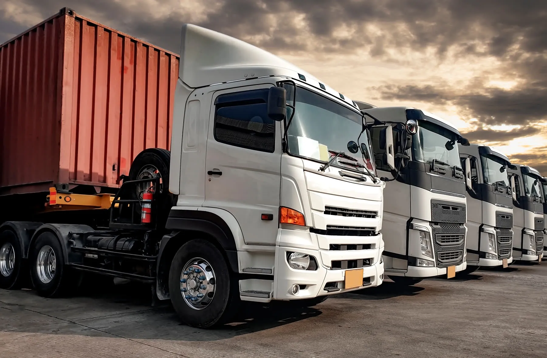 A row of semi-trucks with a shipping container, parked under a dramatic sky at sunset
